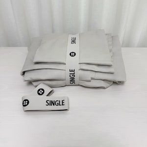 Könnte beinhalten: Ein Stapel hellgrauer Bettwäsche, gesichert mit einem weißen Band mit dem schwarzen Aufdruck "SINGLE". Das Band hat auch ein schwarzes kreisförmiges Logo. Zwei zusätzliche Bänder mit dem gleichen Design befinden sich davor.
