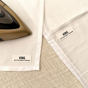 May include: Two white fabric labels with black text that reads "KING BOTTOM RIGHT CORNER" on a white fabric surface. An iron is visible in the background.