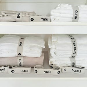 May include: White bed linens neatly folded and organized on shelves, secured with light gray bands labeled with bed sizes: Single, Twin, Queen, Full, Double, Super King, and King. The bands have a black button detail.