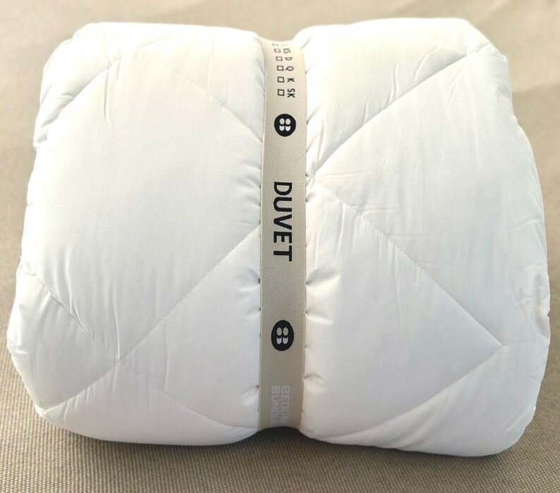 May include: A rolled-up white duvet with a diamond-stitched pattern. The duvet is secured with a light gray band that reads "DUVET" in black text. The band also features two button graphics.