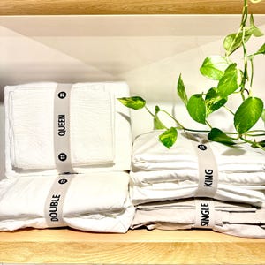 May include: White bed sheet sets, neatly stacked on a wooden shelf. Each set is secured with a gray band labeled with bed sizes: Queen, Double, King, and Single. A green plant with vibrant leaves adds a natural touch.