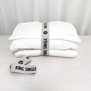 May include: Three white linen pillowcases stacked on top of each other. The pillowcases are tied together with a white elastic band with the text "KING SINGLE" printed in black.