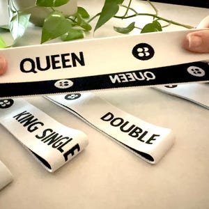 May include: White fabric labels with black text that reads "QUEEN", "DOUBLE", and "KING SINGLE". The labels have a black border and a small graphic. The labels are being held up in front of a green plant.