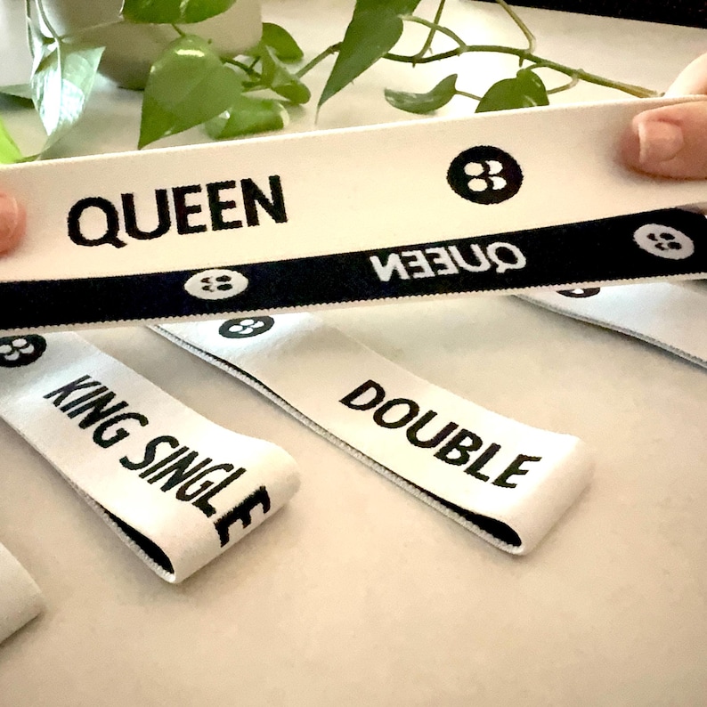 May include: White fabric labels with black text that reads "QUEEN", "DOUBLE", and "KING SINGLE". The labels have a black border and a small graphic. The labels are being held up in front of a green plant.