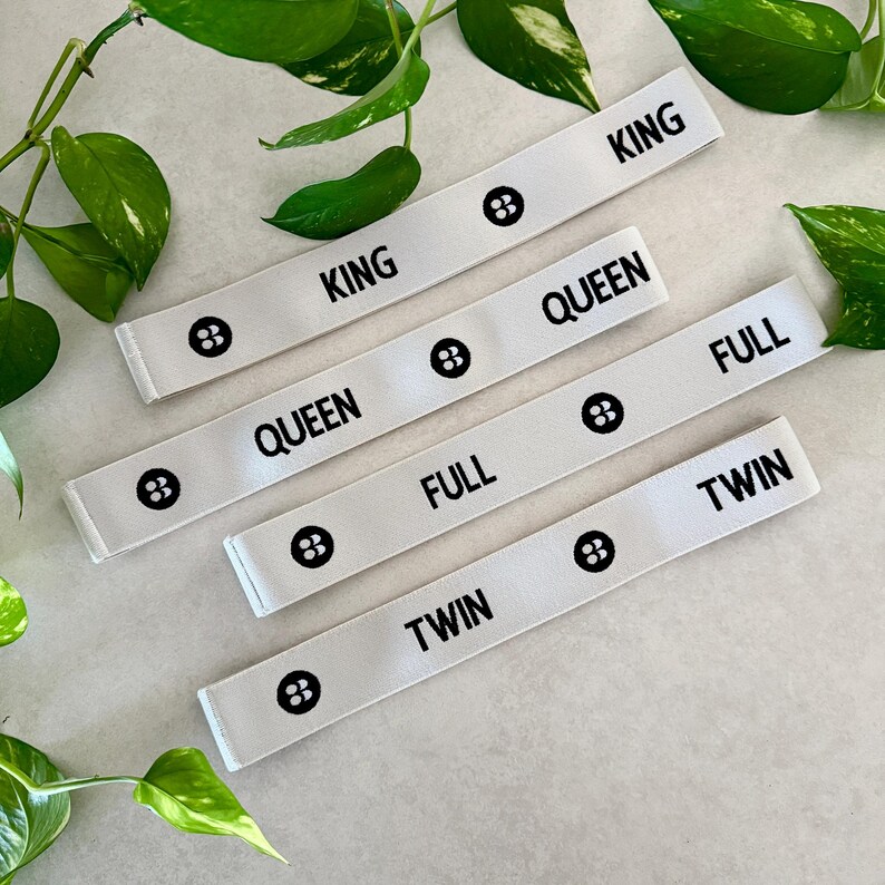 May include: White fabric bed size labels with black text that reads "KING", "QUEEN", "FULL", and "TWIN". Each label has a black graphic. Green leaves are in the background.