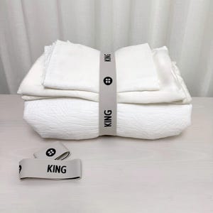 Pode incluir: Conjunto de roupa de cama branco tamanho king, dobrado e amarrado com uma faixa elástica branca com a palavra "KING" impressa. A faixa tem um detalhe de botão.