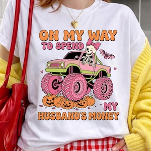 Op de afbeelding: Een wit t-shirt met een afbeelding van een skelet met een roze strik die in een roze monstertruck over pompoenen rijdt. De tekst luidt ON MY WAY TO SPEND MY HUSBAND'S MONEY. Een kleine vleermuis en roze hartjes omringen de illustratie.