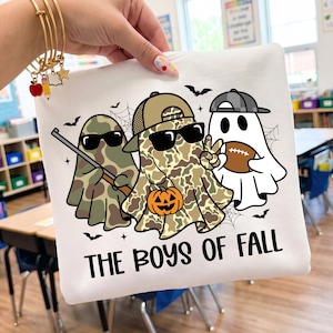 May include: A white sweatshirt featuring a graphic of three ghosts wearing hats and sunglasses. One ghost holds a rifle, one holds a pumpkin, and one holds a football. The text "THE BOYS OF FALL" is printed in black below the ghosts.