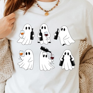May include: White t-shirt featuring a playful design of cartoon ghosts holding wine glasses and bottles. The ghosts are depicted in black and white, with wine glasses filled with red and orange liquid. A fun, whimsical design for Halloween or wine lovers.