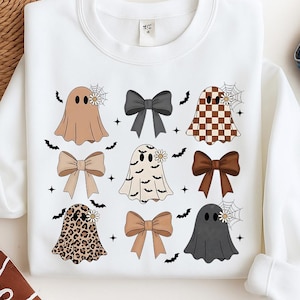 Op de afbeelding: Witte sweatshirt met een patroon van cartoongeesten in verschillende kleuren en patronen, waaronder bruin, geblokt en luipaardprint. Elke geest heeft een madeliefje en spinnenwebdetail. Zwarte strikken en vleermuizen zijn ook afgebeeld.