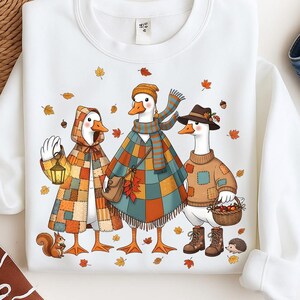 May include: White sweatshirt with a charming illustration of three geese in autumnal outfits. One goose is in a patchwork cape and holds a lantern, another in a plaid poncho and scarf, and the third in a sweater and hat, carrying a fruit basket.