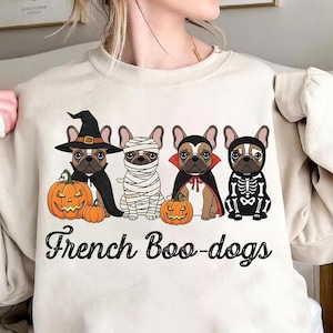 May include: Beige sweatshirt featuring a Halloween-themed graphic. The design includes four French Bulldogs dressed in costumes: a witch, a mummy, a vampire, and a skeleton. Each dog is paired with a carved pumpkin. The text "French Boo-dogs" is below the dogs.