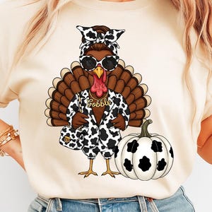 May include: A beige t-shirt featuring a cartoon turkey wearing sunglasses, a cow-print jacket, and a matching purse. The turkey has a gold necklace that says "Gobble" and is next to a white pumpkin with black spots. The turkey is wearing a cow-print headband.
