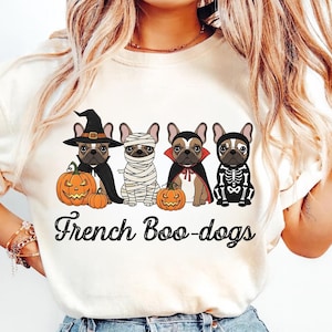 May include: Cream-colored t-shirt featuring a Halloween-themed graphic. The design includes four French Bulldogs dressed in costumes: a witch, a mummy, a vampire, and a skeleton. The phrase "French Boo-dogs" is written in a script font below the dogs.