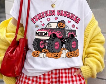 Puede incluir: Una camiseta blanca con el gráfico de un camión monstruo negro con llamas rosas, conducido por una calabaza con gorra. El texto PUMPKIN CRUSHER aparece arriba, rodeado de corazones rosas, flores y calabazas naranjas en la parte inferior.