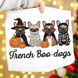 May include: White Halloween-themed graphic featuring four French Bulldogs in costumes: a witch, mummy, vampire, and skeleton. Each dog is next to a carved pumpkin. The text "French Boo-dogs" is below the dogs.
