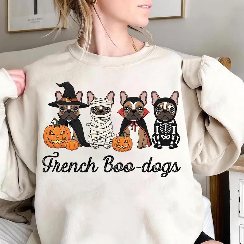 May include: Beige sweatshirt featuring a Halloween-themed graphic. The design includes four French Bulldogs dressed in costumes: a witch, a mummy, a vampire, and a skeleton. Each dog is paired with a carved pumpkin. The text "French Boo-dogs" is below the dogs.