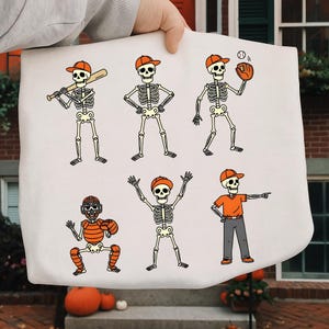 May include: A white tote bag featuring six cartoon skeletons in baseball attire. The skeletons are wearing orange caps and are in various baseball poses, including batting, pitching, and catching. The design is outlined in black.