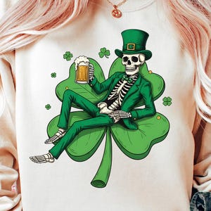 Op de afbeelding: Een witte sweatshirt met een skelet in een groen pak en hoge hoed, met een biermok. Het skelet zit op een grote groene klaver, met kleinere klavers eromheen. Een ontwerp met een St. Patrick's Day-thema.