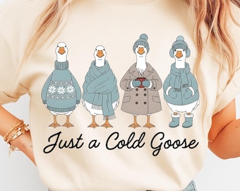 Puede incluir: Camiseta color crema con cuatro gansos de dibujos animados, cada uno vestido con ropa de invierno. Los gansos llevan suéteres, bufandas, gorros y abrigos. El texto "Just a Cold Goose" está escrito en cursiva debajo.