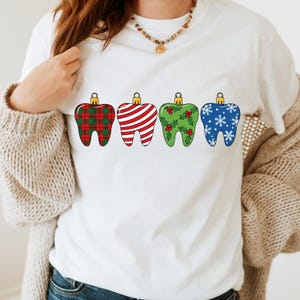 Puede incluir: Camiseta blanca con cuatro adornos dentales estilizados. Cada diente est&aacute; decorado con un patr&oacute;n festivo diferente: cuadros rojos, rayas rojas y blancas, acebo verde y copos de nieve azules. Los adornos tienen tapas doradas.