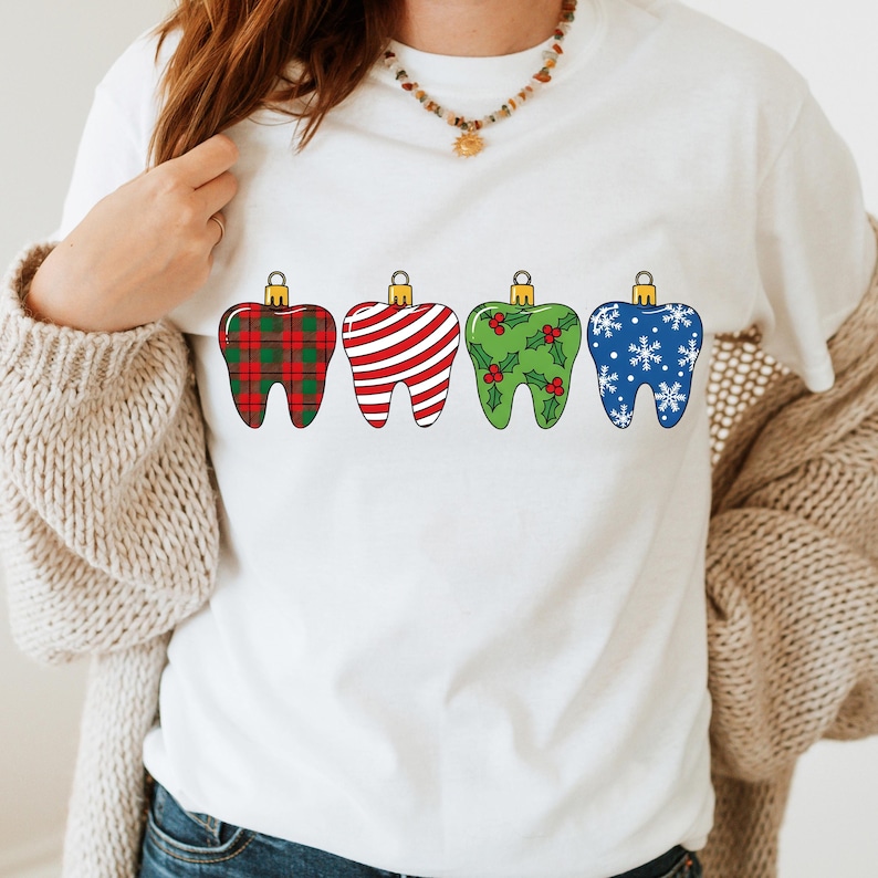 Puede incluir: Camiseta blanca con cuatro adornos dentales estilizados. Cada diente est&aacute; decorado con un patr&oacute;n festivo diferente: cuadros rojos, rayas rojas y blancas, acebo verde y copos de nieve azules. Los adornos tienen tapas doradas.