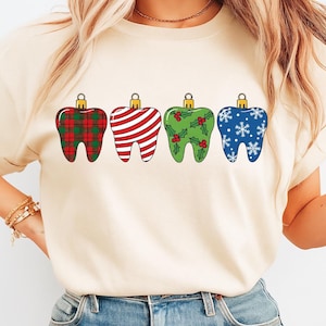 Puede incluir: Una camiseta color crema con cuatro adornos navide&ntilde;os en forma de diente. Los adornos est&aacute;n decorados con un estampado de cuadros rojos y verdes, rayas rojas y blancas, acebo verde y copos de nieve azules. Cada adorno tiene una tapa dorada.