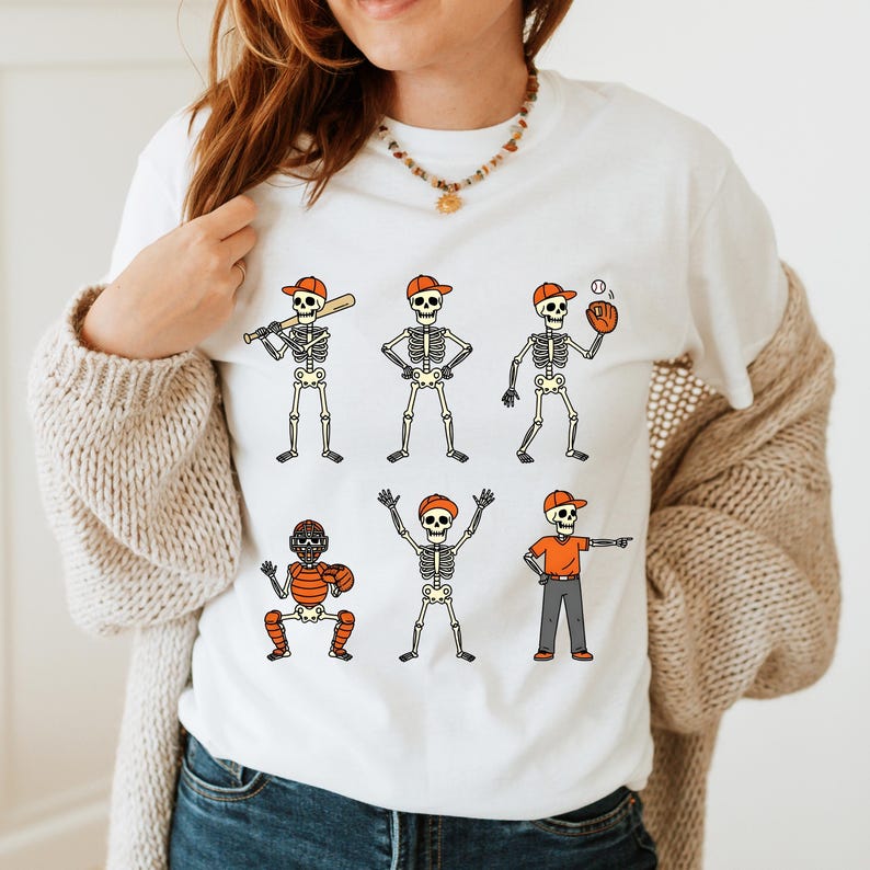 May include: White t-shirt featuring a baseball-themed skeleton design. The design includes six cartoon skeletons in various baseball positions, each wearing an orange cap. The skeletons are in different poses, holding a bat, ball, and glove.