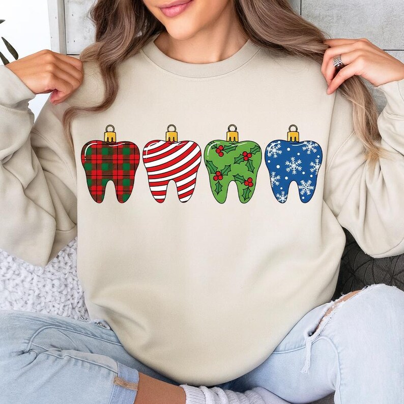 Puede incluir: Una sudadera beige con cuatro adornos navide&ntilde;os en forma de diente. Los adornos est&aacute;n decorados con un estampado de cuadros rojos y verdes, rayas rojas y blancas, acebo y bayas verdes y copos de nieve azules. Cada adorno tiene una tapa dorada.