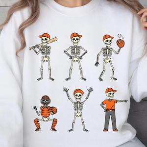 May include: White sweatshirt featuring six cartoon skeletons in baseball attire. The skeletons wear orange caps and are depicted batting, pitching, catching, and in other baseball poses. The design is outlined in black.