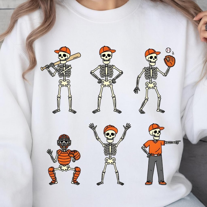 May include: White sweatshirt featuring six cartoon skeletons in baseball attire. The skeletons wear orange caps and are depicted batting, pitching, catching, and in other baseball poses. The design is outlined in black.