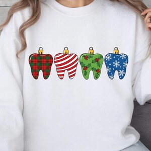 Puede incluir: Sudadera blanca con cuatro dise&ntilde;os de adornos navide&ntilde;os en forma de diente. Los adornos presentan diferentes patrones: cuadros rojos, rayas rojas y blancas, verde con acebo y azul con copos de nieve. Cada adorno tiene una tapa dorada.