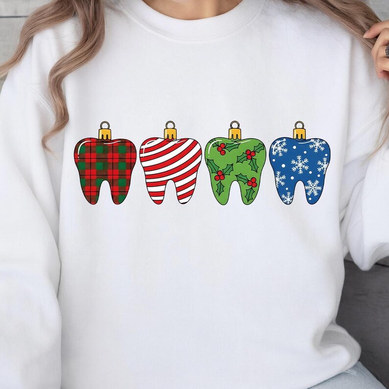 Puede incluir: Sudadera blanca con cuatro dise&ntilde;os de adornos navide&ntilde;os en forma de diente. Los adornos presentan diferentes patrones: cuadros rojos, rayas rojas y blancas, verde con acebo y azul con copos de nieve. Cada adorno tiene una tapa dorada.