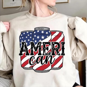 May include: A cream-colored sweatshirt featuring a graphic of three stylized beverage cans. The cans are decorated with red and white stripes and blue fields of white stars, with the word "AMERICAN" in bold black letters.