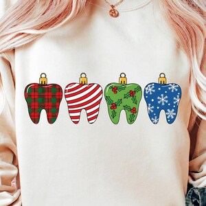 Puede incluir: Una sudadera crema con cuatro adornos navide&ntilde;os en forma de diente. Los adornos est&aacute;n decorados con un estampado de cuadros rojos y verdes, rayas rojas y blancas, acebo verde y copos de nieve azules. Cada adorno tiene una tapa dorada.