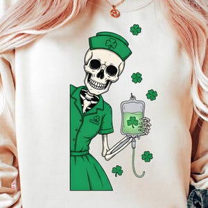 Op de afbeelding: Een witte sweatshirt met een cartoon skelet in een groene verpleegstersuniform en pet, met een zak groene vloeistof met een klavertje vier. Groene klavers zijn verspreid over de afbeelding.