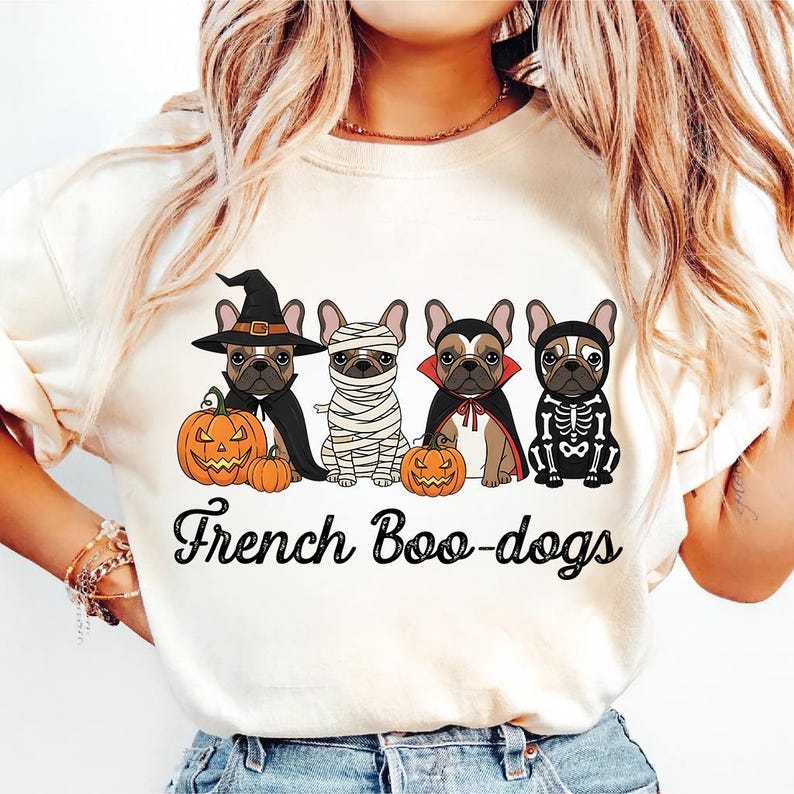 May include: Cream-colored t-shirt featuring a Halloween-themed graphic. The design includes four French Bulldogs dressed in costumes: a witch, a mummy, a vampire, and a skeleton. The phrase "French Boo-dogs" is written in a script font below the dogs.