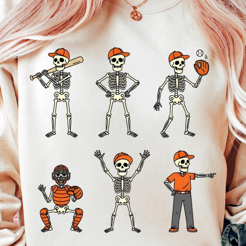 May include: Cream-colored sweatshirt featuring a baseball-themed skeleton design. Six cartoon skeletons are depicted in various baseball positions, wearing orange caps and uniforms. One holds a bat, another a glove, and a third is catching.