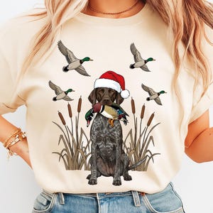 May include: A beige t-shirt featuring a German Shorthaired Pointer wearing a Santa hat and holding a duck in its mouth. Ducks are flying in the background. Cattails are in the foreground.