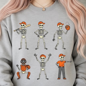 May include: A light gray sweatshirt featuring a baseball-themed skeleton design. The graphic includes six skeletons in various baseball poses, each wearing an orange baseball cap. One skeleton holds a bat, another a glove, and another is catching.