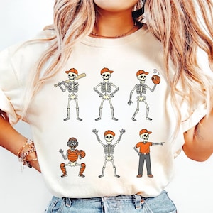 May include: Cream-colored sweatshirt featuring six cartoon skeletons in baseball poses. Each skeleton wears an orange baseball cap and is engaged in different baseball activities, such as batting, pitching, and catching.