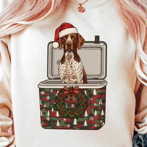 Puede incluir: Una sudadera blanca con un Braco Alemán con un gorro de Papá Noel, dentro de una nevera. La nevera tiene un patrón de camuflaje con copos de nieve y árboles de Navidad, y una corona con un lazo rojo.