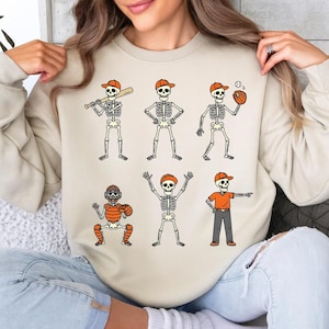May include: Beige sweatshirt featuring six baseball-themed skeleton illustrations. Each skeleton wears an orange baseball cap and is engaged in different baseball actions, such as batting, catching, and pitching. The design is playful and suitable for sports fans.
