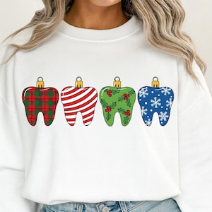 Puede incluir: Sudadera blanca con cuatro adornos navide&ntilde;os en forma de diente. Los adornos tienen varios patrones: cuadros rojos, rayas rojas y blancas, verde con acebo y azul con copos de nieve. Cada adorno tiene una tapa dorada.