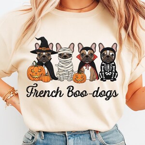 May include: Beige t-shirt featuring four French Bulldogs dressed in Halloween costumes: a witch, mummy, vampire, and skeleton. Each dog is next to a carved pumpkin. The text "French Boo-dogs" is printed below the dogs.