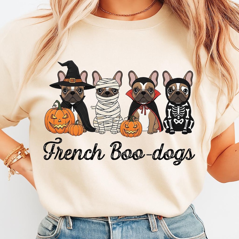 May include: Beige t-shirt featuring four French Bulldogs dressed in Halloween costumes: a witch, mummy, vampire, and skeleton. Each dog is next to a carved pumpkin. The text "French Boo-dogs" is printed below the dogs.