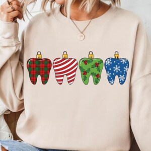 Puede incluir: Una sudadera beige con cuatro adornos navide&ntilde;os en forma de diente. Los adornos tienen diferentes patrones: cuadros rojos y verdes, rayas rojas y blancas, verde con acebo y azul con copos de nieve. Cada uno tiene una tapa dorada.