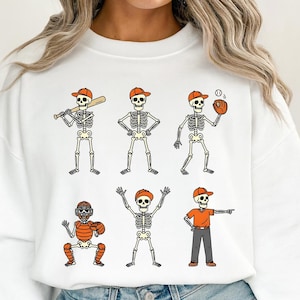 May include: White sweatshirt featuring six skeleton baseball players in orange caps and uniforms. The skeletons are in various baseball positions, including batting, pitching, and catching. A fun, sporty design for Halloween or baseball fans.