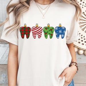 Puede incluir: Una camiseta color crema con cuatro dise&ntilde;os de dientes decorativos. Cada diente est&aacute; dise&ntilde;ado como un adorno navide&ntilde;o, con patrones que incluyen cuadros escoceses, rayas, acebo y copos de nieve. Los dientes tienen tapas doradas.
