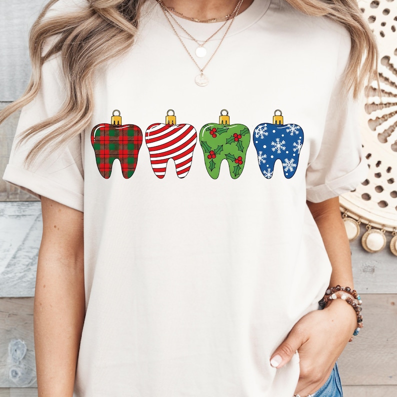 Puede incluir: Una camiseta color crema con cuatro dise&ntilde;os de dientes decorativos. Cada diente est&aacute; dise&ntilde;ado como un adorno navide&ntilde;o, con patrones que incluyen cuadros escoceses, rayas, acebo y copos de nieve. Los dientes tienen tapas doradas.